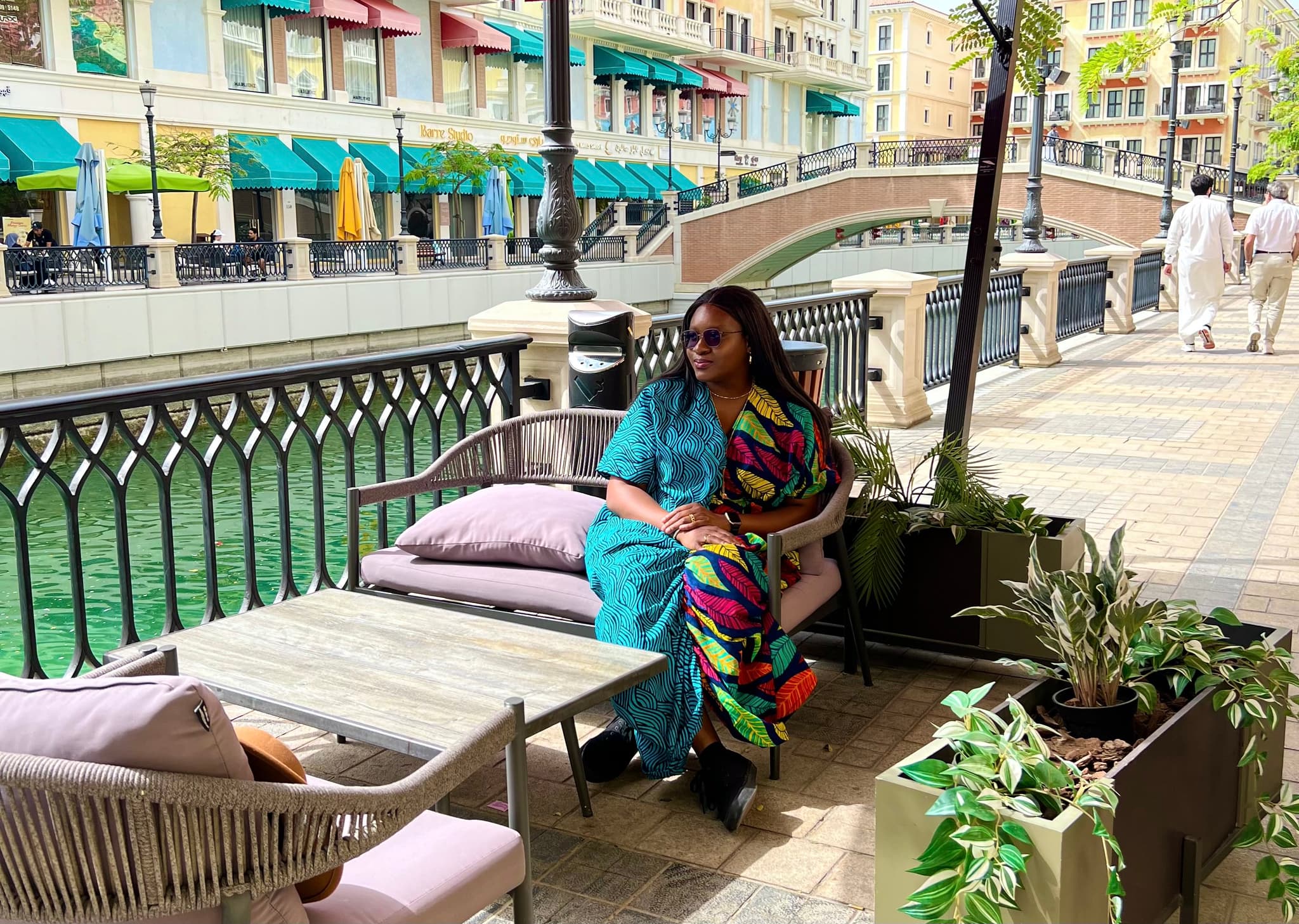 A woman sits by a canal in a colorful outfit, with pastel buildings and a small bridge in the background.